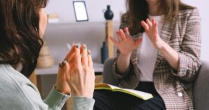 Two women in a counseling session; one gestures with hands while the other sits with an open notebook on her lap.