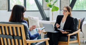Therapist with glasses and a laptop meets with a client holding a smartphone in a bright, modern office.