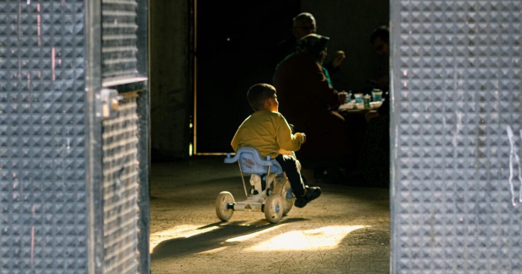 Child in a yellow shirt rides a small tricycle through a doorway toward a sunlit room where adults sit at a table in the background.