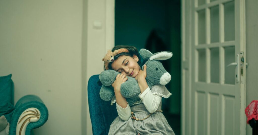 Young girl wearing a headband with a small animal, hugging a gray stuffed donkey while sitting in a doorway indoors.