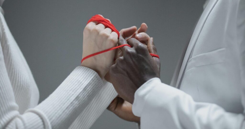 image - Dallas Mental Health Two people hold hands with a red thread wrapped around their wrists, symbolizing unity and connection.